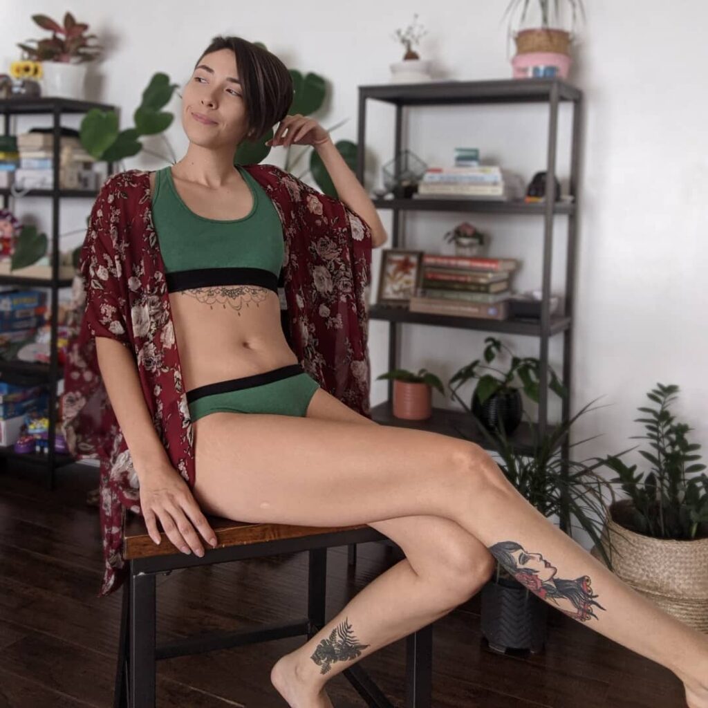 A-tattooed-woman-wearing-a-green-bralette-green-underwear-and-a-red-floral-robe-sits-on-a-chair-in-a-room-full-of-bookshelves-and-plants